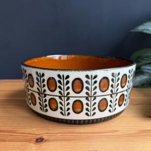 May include: A round ceramic bowl featuring a white exterior with a repeating pattern of dark green vertical leaf stems and orange oval shapes. The interior of the bowl is a solid, glossy orange colour, and the base has a dark brown ribbed texture.