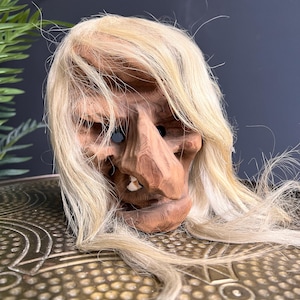 Puede incluir: Una máscara de madera tallada que representa un rostro anciano con una nariz larga, un solo diente y cabello rubio. La máscara tiene cuencas oscuras y probablemente sea una pieza decorativa o teatral.