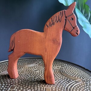 May include: A wooden horse figurine, reddish-brown in colour, with carved details such as a mane and bridle. The horse stands on a decorative surface, displaying the wood grain. The figurine is a charming example of handcrafted wooden art.