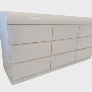 May include: A white, modern dresser with nine drawers. The dresser has a curved top and a glossy finish. The drawers are arranged in three rows of three. The dresser is a light neutral color and sits on a low base.