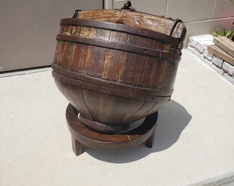 May include: A large, round, wooden bucket with metal bands sits on a wooden stand. The bucket is brown and has a worn, rustic appearance.