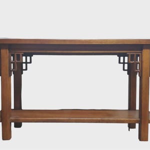 May include: A wooden console table with a warm brown finish. The table features a rectangular top, a lower shelf, and decorative geometric cutouts on the legs. The table's design suggests an Asian-inspired aesthetic.