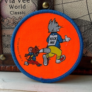 Op de afbeelding: Een ronde geborduurde patch met een feloranje achtergrond en een blauwe rand. Het toont een cartoonkat in een blauw shirt met een wit bordje op de rug met de tekst KICK ME, terwijl een bruine muis aan de staart van de kat trekt. 1990 Turner Ent. Co.