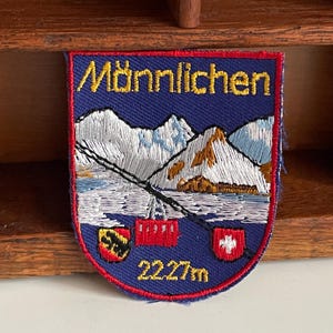 May include: Embroidered patch with a blue background and a red border. The patch features the word "Männlichen" in yellow, along with a mountain scene, a cable car, and the text "2227m". Includes two crests.