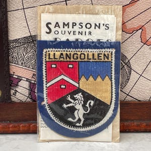Peut inclure: Un écusson souvenir dans une pochette en plastique transparent avec une carte d'en-tête portant l'inscription SAMPSON'S SOUVENIR BADGES. L'écusson en forme de blason affiche le texte LLANGOLLEN au-dessus d'un motif rouge, bleu et noir.