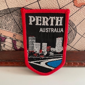Op de afbeelding: Een zwart en rood embleem met de tekst "PERTH AUSTRALIA" in zilver. Het embleem toont een skyline-illustratie met een blauw waterelement en rode accenten. Het embleem heeft een rode rand.