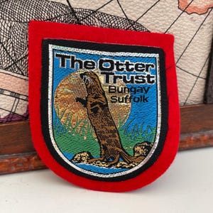 May include: A shield-shaped embroidered patch with a bright red felt border. The centre features a brown otter standing against a gold circular background, with blue water, green grass, and the text The Otter Trust Bungay Suffolk in black.