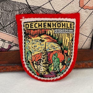 May include: A red, shield-shaped patch with the words "DECHENHOHLE" and "KAISERHALLE" at the top. The patch features a colourful landscape scene with a tower and trees. The patch has a white border.
