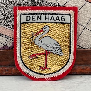 May include: A shield-shaped embroidered patch featuring a white stork with a red beak and legs on a gold background. The top section contains the text DEN HAAG in black letters on a white field. The entire patch is bordered by a thick red edge.
