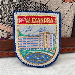 May include: Embroidered patch with a blue border, featuring the text "hotel ALEXANDRA" in gold lettering on a red background. The patch depicts a building, mountains, and the text "LOEN NORWAY".