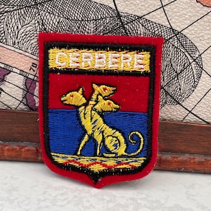 Puede incluir: Parche bordado con forma de escudo. El parche presenta un fondo rojo con una franja azul y un perro amarillo de tres cabezas. La palabra "CERBERE" está bordada en blanco en la parte superior.