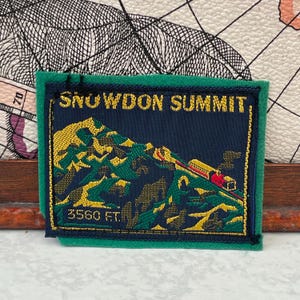 以下が含まれることがあります： 緑色のフェルトの縁取りが付いた長方形の刺繍ワッペン。濃い青色の中心部には、黄色の山のグラフィック、線路上の赤と黄色の列車、そして黄色の大きな文字で「SNOWDON SUMMIT 3560 FT.」と記されています。
