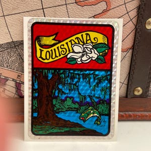 May include: A sticker with a two-part design. The top section has a red background with a yellow banner reading "Louisiana" and a white magnolia flower. The bottom section depicts a swamp scene with a jumping fish.