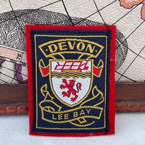Puede incluir: Un parche rectangular con borde rojo y centro azul marino. El parche presenta la palabra "DEVON" en letras doradas en la parte superior y "LEE BAY" en la parte inferior. Un escudo con un león rojo está en el centro.
