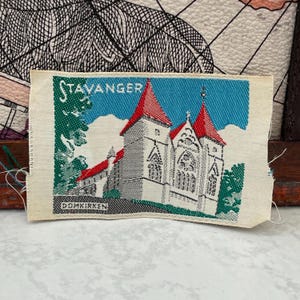May include: A rectangular woven fabric patch featuring a detailed illustration of the Stavanger Domkirken cathedral. The design includes red roofs, white stone walls, and green foliage against a blue sky, with the text STAVANGER and DOMKIRKEN.