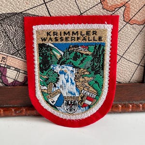 Könnte beinhalten: Ein roter, schildförmiger Aufnäher mit einem detaillierten Stickdesign. Das Design zeigt einen Wasserfall, Berge und den Text "KRIMMLER WASSERFALLE" und "AUSTRIA". Der Aufnäher ist mit weißen und goldenen Nähten eingefasst.