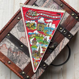 Könnte beinhalten: Ein rotes dreieckiges Wimpel mit dem Wort "ÖSTERREICH" und Illustrationen österreichischer Städte. Der Wimpel hat weiße und goldene Verzierungen und den Text "GRUSS AUS". Er wird auf einer Vintage-Holzkiste präsentiert.