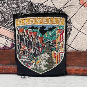 May include: A rectangular embroidered patch with a black border featuring a woven scene of a village with red-roofed buildings, green trees, and a path. The word CLOVELLY is stitched in gold thread across the top section of the patch design.