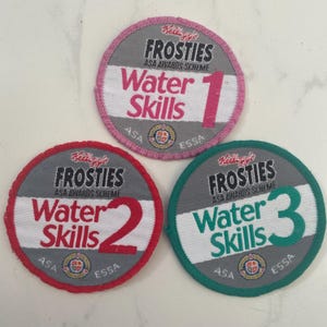 May include: Three round fabric patches with the text "Kellogg's FROSTIES ASA AWARDS SCHEME Water Skills" and numbers 1, 2, and 3. Each patch has a different colored border: pink, red, and green. The patches are gray, white, and feature the ASA and ESSA logos.