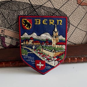 May include: Embroidered patch featuring the city of Bern, Switzerland. The patch has a blue background with the word "BERN" in gold letters. The design includes a bear, a cityscape, and the Swiss flag.