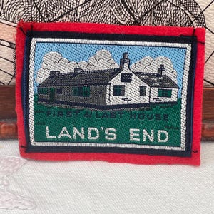 Może przedstawiać: Prostokątna naszywka z czerwoną obwódką. Haftowany obraz przedstawia budynek z ciemnym dachem, zieloną trawą i napisem "FIRST & LAST HOUSE LAND'S END". Niebo jest jasnoniebieskie z białymi chmurami. Pamiątka.