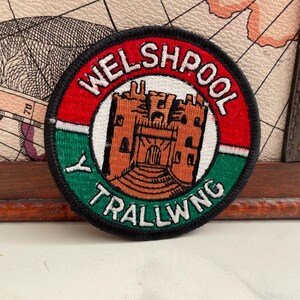 May include: A circular patch with the text WELSHPOOL and TRALLWNG. The patch features a brown castle illustration in the centre, surrounded by red, white, green, and black borders. It is likely a souvenir or emblem.