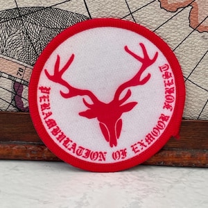 Op de afbeelding: Een ronde geborduurde patch met het silhouet van een rode hertenkop in het midden op een witte achtergrond. De rand is rood en de tekst "PERAMBULATION OF EXMOOR FOREST" is in rode letters langs de gebogen rand van de patch geborduurd.