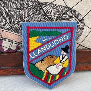 May include: Embroidered patch with a blue background and red border. The patch features the word "LLANDUDNO" and a colourful illustration of a landscape with a figure in traditional clothing. The patch is likely a souvenir or emblem.