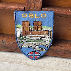 May include: Embroidered patch with the word "OSLO" in gold lettering. The patch depicts a detailed illustration of buildings and a harbour scene, set against a blue backdrop. The shield-shaped patch includes a Norwegian flag design at the base.