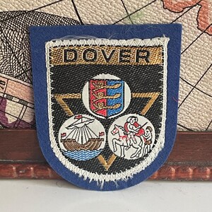May include: A blue patch with the word "DOVER" at the top. The patch features a shield with a coat of arms, a ship, and a knight on horseback. The patch is trimmed with white stitching.