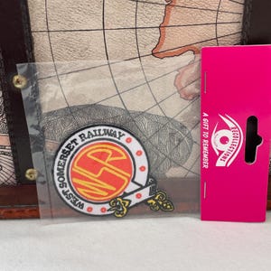 May include: A West Somerset Railway patch with a red and yellow emblem, displayed in a clear plastic sleeve. The patch is on a background of a map design. A pink gift tag with the text "A Gift to Remember" is on the right.