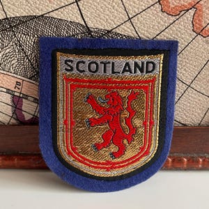 May include: A blue and gold embroidered patch featuring the word "SCOTLAND" above a red lion rampant emblem. The patch has a shield shape with a blue border and gold accents.