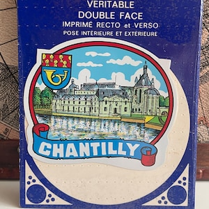 May include: A blue box with the words "VERITABLE DOUBLE FACE" and other French text. The box features a circular design with a detailed illustration of a building on a lake, with the word "CHANTILLY" in a banner below.