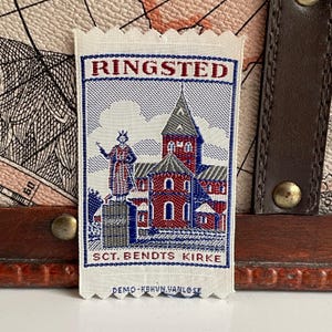 May include: A rectangular fabric patch with the word "RINGSTED" in red letters at the top. Below is an illustration of a church and a statue, with the text "SCT. BENDTS KIRKE" at the bottom. The patch is white with blue and red details.