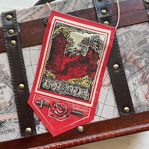 May include: A red and white fabric banner with the word "Furanda" printed in black. The banner features a vintage illustration of a castle with a red roof and a green tree. A red rose and a sword are depicted at the bottom of the banner.