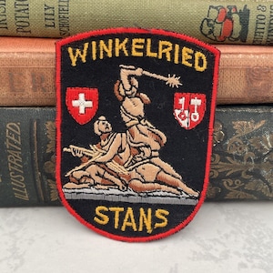 May include: A black embroidered patch with a red border. The patch features a scene of a man holding a spear, standing over two other figures. The text "WINKELRIED" is at the top of the patch, and "STANS" is at the bottom. The patch also features two shields, one with a red cross on a white background and the other with a red cross on a white background with a hammer and anvil.