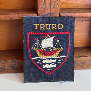 May include: A rectangular navy blue embroidered patch featuring the word TRURO in gold thread above a shield. The shield contains a gold ship with white sails on stylized white waves, all outlined in red thread on a dark blue background.