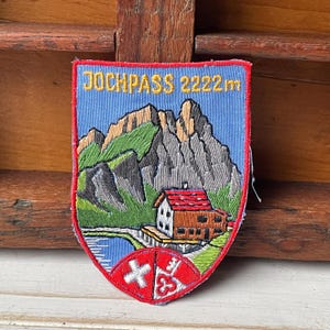 May include: Embroidered patch with a shield shape, featuring a mountain scene with a cabin, bridge, and lake. The text "JOCHPASS 2222m" is at the top. The bottom has a red and white emblem. The patch has a blue background.