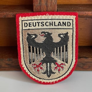 May include: A shield-shaped embroidered patch featuring a black eagle with red claws and beak on a gold background. The word DEUTSCHLAND is stitched in black capital letters on a cream-colored horizontal band at the top, bordered by red felt.