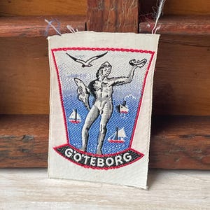 May include: A rectangular woven patch with a design of a nude figure holding a fish and a dish. The patch has a red border and the word "GOTEBORG" at the bottom. Two seagulls and sailboats are also depicted.