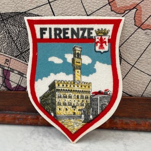 以下が含まれることがあります： 黒い文字で「FIRENZE」と書かれた盾形の刺繍ワッペン。黄色い建物と高い塔が、白い雲が浮かぶ水色の空を背景に描かれています。右上には、赤いフルール・ド・リスが描かれた小さな白い紋章があしらわれています。