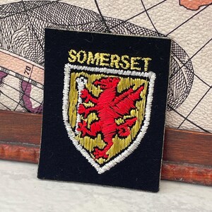 Puede incluir: Parche bordado negro con la palabra "SOMERSET" en letras doradas. Presenta un emblema de dragón rojo sobre un escudo dorado y blanco. El parche es rectangular.