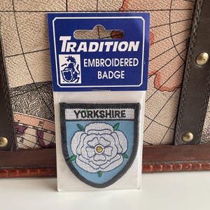 May include: An embroidered badge featuring the word "Yorkshire" above a white rose on a light blue background. The badge is in a clear plastic package with a blue and white header that reads "Tradition Embroidered Badge".