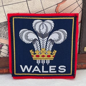 May include: A square patch with a navy blue background, featuring a gold-colored crown and three white feathers. The word "WALES" is displayed in white letters at the bottom. The patch has a red border and a gold trim.