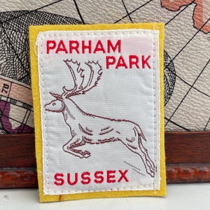 May include: A rectangular patch with a white centre and yellow border. The patch features the words "PARHAM PARK SUSSEX" in red, with a detailed illustration of a deer.