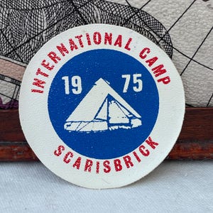 May include: A round, off-white patch with a blue circle in the center. The text "INTERNATIONAL CAMP" and "SCARISBRICK" are in red around the edge. Inside the blue circle are the numbers "19 75" and a white tent graphic.