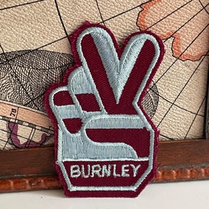 Puede incluir: Un parche bordado en color granate y azul claro que muestra una mano haciendo el signo de la paz. El diseño incluye la palabra BURNLEY en letras mayúsculas de color azul claro en la parte inferior, con un borde granate alrededor de la mano.