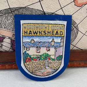 May include: A blue fabric patch with a white border. The patch features a detailed image of a building with the text "WORDSWORTH'S LODGING HAWKSHEAD" at the top. The image depicts a stone building with a blue roof and green foliage.