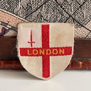 Puede incluir: Un parche en forma de escudo con fondo crema, que presenta una cruz roja y la palabra "LONDON" en letras doradas. Una espada roja está encima de la cruz. El parche es probablemente un recuerdo o un emblema.