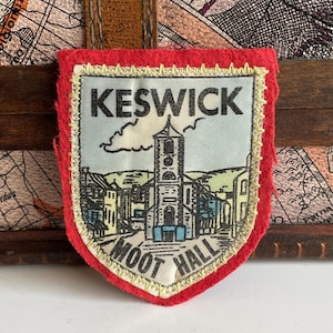 Puede incluir: Un parche de fieltro rojo con un borde bordado en oro. El parche presenta una ilustración vintage de una ciudad con una iglesia y el texto "KESWICK MOOT HALL".